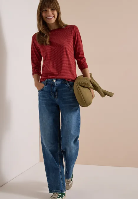 Langarm Shirt mit Boat Neck und Streifen russet red Eine junge Frau in rotem Langarmshirt und weiten Jeans hält ein grünes Cap und lächelt.