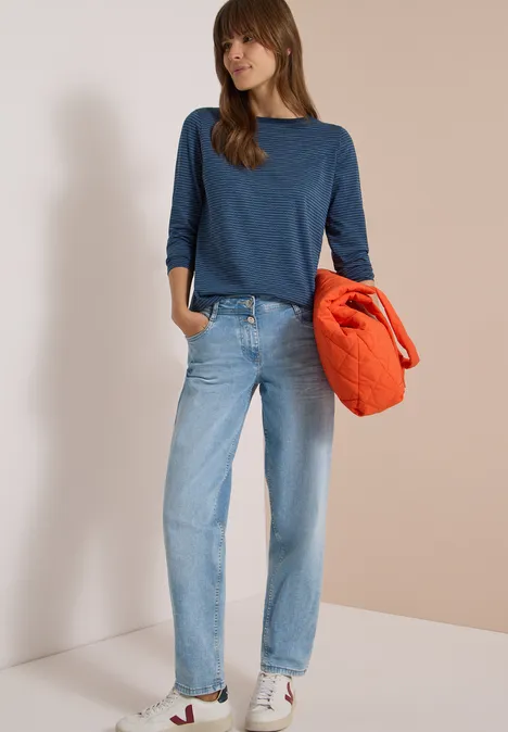 Langarm Shirt mit Boat Neck und Streifen moonlight blue Frau in Jeans, blauem Pullover und weißen Sneakern hält eine orange Jacke vor neutralem Hintergrund.