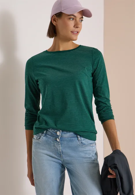 Langarm Shirt mit Boat Neck und Streifen dark fir green Frau mit grünem Langarmshirt, Jeans, Cap und Jacke in der Hand vor zweifarbigem Hintergrund.