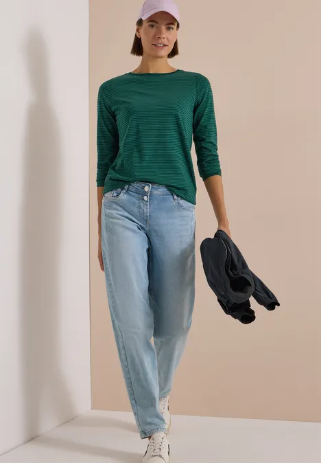 Langarm Shirt mit Boat Neck und Streifen dark fir green Frau in grünem Dreiviertelshirt, Jeans und Cap hält Jacke und geht vor schlichtem Hintergrund.
