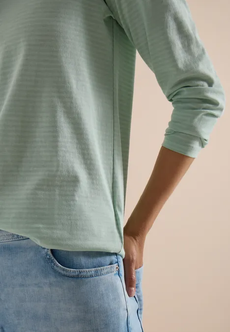 Person trägt hellgrünes Langarmshirt und Jeans, Hand steckt lässig in Gesäßtasche.
