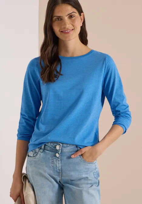 Vrouw in blauw langarmshirt en lichtblauwe jeans, lacht met hand in de zak.