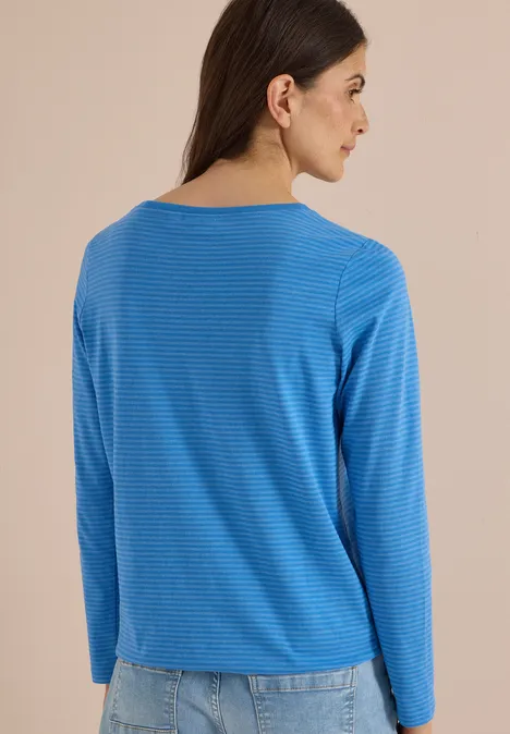Vrouw van achteren in blauw gestreept shirt met lange mouwen en spijkerbroek, beige achtergrond.