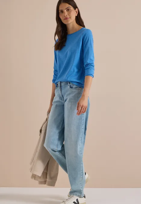 Vrouw in blauw shirt met lange mouwen, wijdere jeans en witte sneakers, houdt lichte jas vast voor neutraal decor.