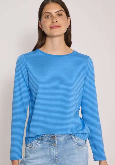 Vrouw in blauw shirt met lange mouwen en spijkerbroek voor een lichte achtergrond.