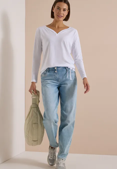 Shirt mit Split Neck im Tunika-Look White Frau in weißem V-Ausschnitt-Shirt, hellblauen Jeans und silbernen Sneakers hält grüne Tasche vor beigem Hintergrund.