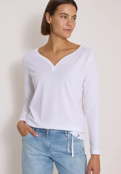 Shirt mit Split Neck im Tunika-Look White Frau in weißem Langarmshirt mit V-Ausschnitt und Jeans, locker posierend vor heller Wand.