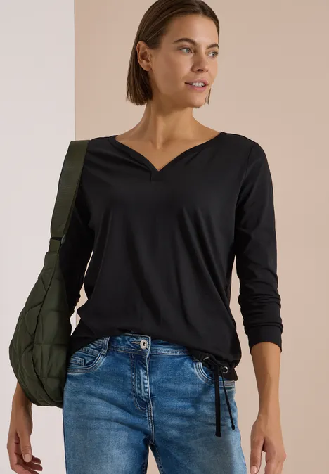 Shirt mit Split Neck im Tunika-Look Black Frau trägt schwarzen V-Ausschnitt-Top mit langen Ärmeln, Jeans und grüner Schultertasche.