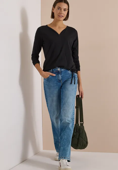 Shirt mit Split Neck im Tunika-Look Black Frau lehnt an Wand, trägt schwarzes V-Shirt, helle Jeans und hält eine grüne Tasche.