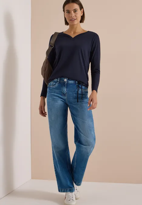 Shirt mit Split Neck im Tunika-Look urban dark blue Frau in weiten Jeans, dunkelblauem Pullover und weißen Sneakern mit Rucksack steht vor hellem Hintergrund.