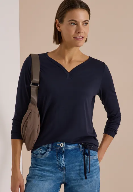 Shirt mit Split Neck im Tunika-Look urban dark blue Frau in dunkelblauem 3/4-Ärmel-Shirt mit V-Ausschnitt, Jeans und Schultertasche, lächelnd seitlich blickend.