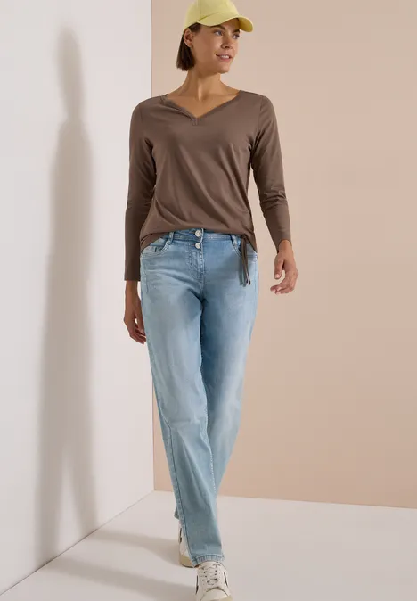 Shirt mit Split Neck im Tunika-Look urban taupe Frau in braunem V-Ausschnittshirt, hellblauer Jeans, weißen Sneakern und gelber Cap, lässig vor beigem Hintergrund.