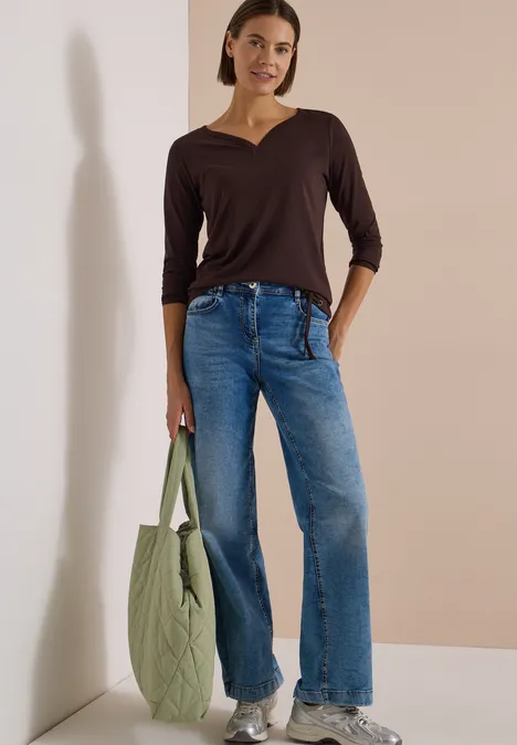 Shirt mit Split Neck im Tunika-Look truffle brown Frau in braunem V-Shirt, weiten Jeans und weißen Sneakers lehnt an hellem Hintergrund und hält eine grüne Tasche.