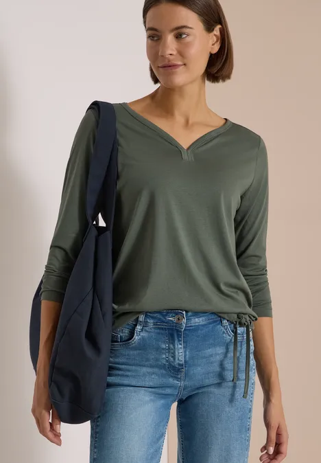 Shirt mit Split Neck im Tunika-Look dried olive green Frau in grüner V-Ausschnitt-Bluse mit 3/4-Ärmeln, blauer Jeans und dunkelblauer Umhängetasche.