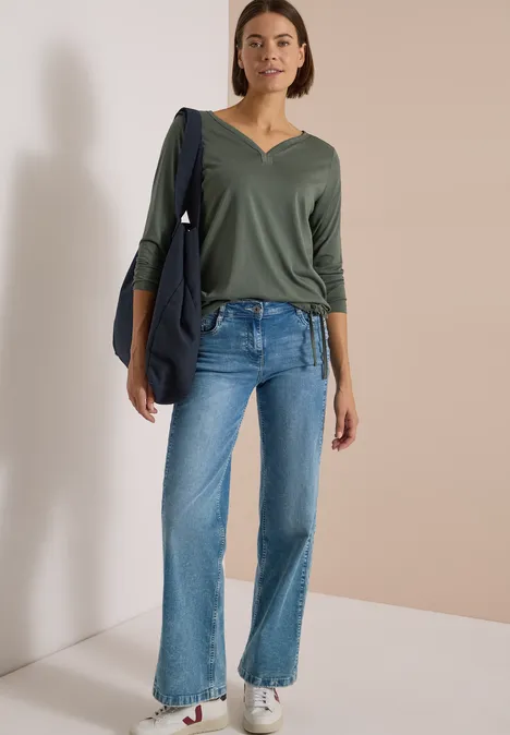 Shirt mit Split Neck im Tunika-Look dried olive green Eine Frau trägt grüne V-Neck-Bluse, blaue Jeans und weiße Sneaker, mit einer dunkelblauen Tasche über der Schulter.