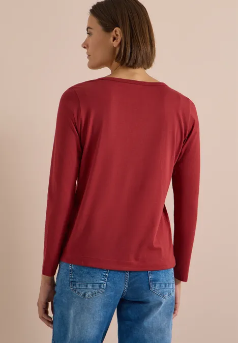 Shirt mit Split Neck im Tunika-Look russet red Rückenansicht einer Person mit rotem Langarmshirt und blauer Jeans vor beigefarbenem Hintergrund.