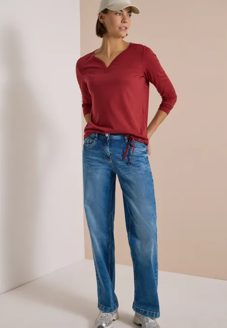 Shirt mit Split Neck im Tunika-Look russet red Frau im roten 3/4-Shirt, blauen Jeans und weißer Baseballkappe vor neutralem Hintergrund.