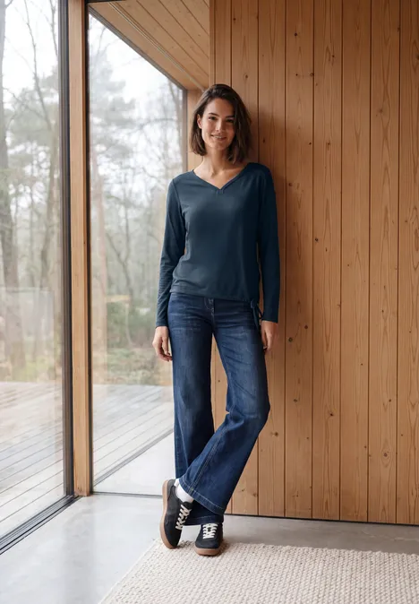 Shirt mit Split Neck im Tunika-Look moonlight blue Lächelnde Frau in Jeans und dunkelblauem Oberteil lehnt entspannt an Holzwand neben Fenster.