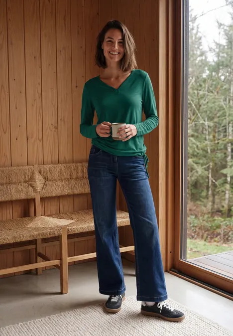 Eine Frau in grüner Bluse und Jeans steht lächelnd mit einer Tasse in einem hellen Holzraum am Fenster.
