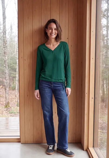 Eine lächelnde Frau in grünem Pullover und Jeans steht vor einer Holzverkleidung neben einem Fenster.