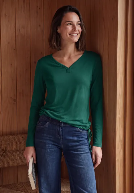 Eine lächelnde Frau in grünem Langarmshirt und Jeans steht in einem Holzraum und hält ein Buch.
