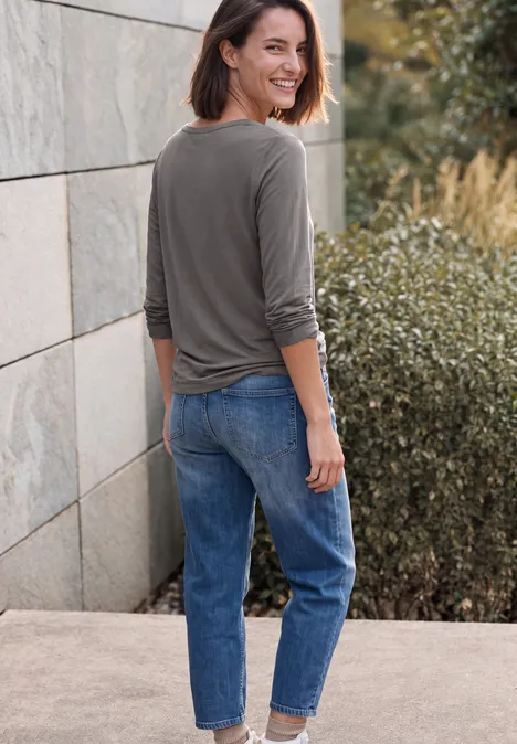 Shirt mit Split Neck im Tunika-Look storm grey Frau steht lächelnd seitlich vor einer Steinmauer, trägt olivgrünes Shirt, blaue Jeans und weiße Sneaker.