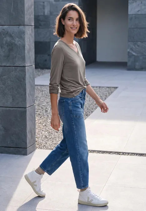 Shirt mit Split Neck im Tunika-Look storm grey Eine junge Frau in Jeans und Sneakern geht lächelnd an modernen Steinpfeilern vorbei.