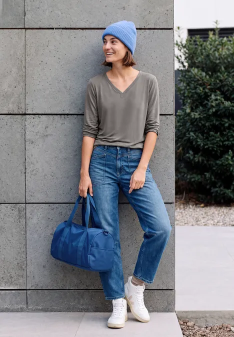 Shirt mit Split Neck im Tunika-Look storm grey Lächelnde Frau in Jeans, grauem Shirt und blauer Mütze lehnt an Wand und hält eine blaue Reisetasche.
