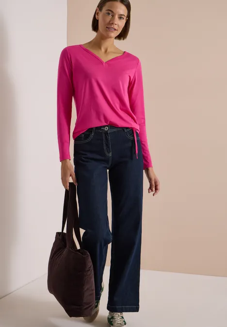 Frau trägt pinken V-Ausschnitt-Pullover, weite Jeans-Hose und hält Tasche vor beigem Hintergrund.