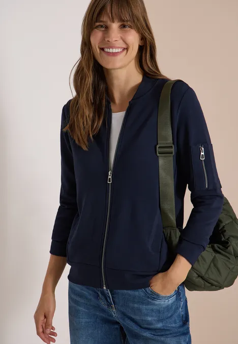 Ottoman Blouson in Unifarbe urban dark blue Eine lächelnde Frau in blauer Strickjacke, Jeans und weißem Shirt, mit grünem Rucksack.