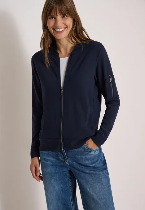 Ottoman Blouson in Unifarbe urban dark blue Lächelnde Frau in dunkelblauer Zip-Jacke und Jeans mit Händen in den Taschen vor weißem Hintergrund.