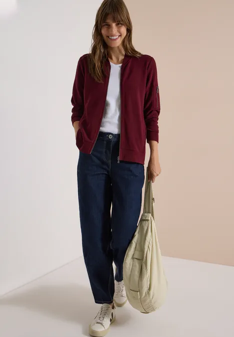Ottoman Blouson in Unifarbe red wine Frau trägt burgunderrote Jacke, weißes Shirt, lockere Jeans, weiße Sneaker und hält eine beige Tasche.