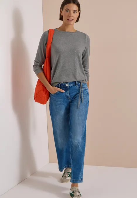 Frau in grauem Pullover, weiten Jeans und roter Tasche vor hellem Hintergrund.