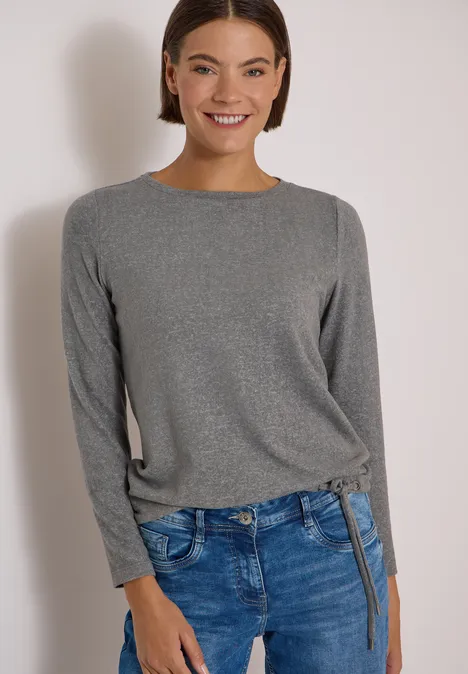 Frau mit grauem Langarmshirt und blauer Jeans, lächelt.