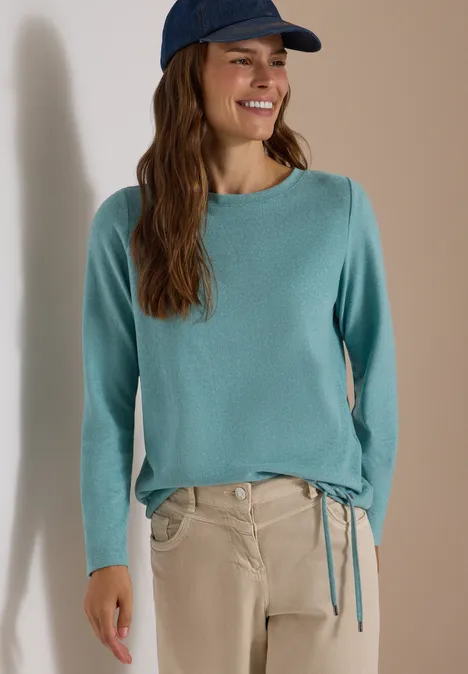 Frau im türkisfarbenen Pullover mit Seitenschnürung, beige Hose und Cap, lächelnd vor neutralem Hintergrund.