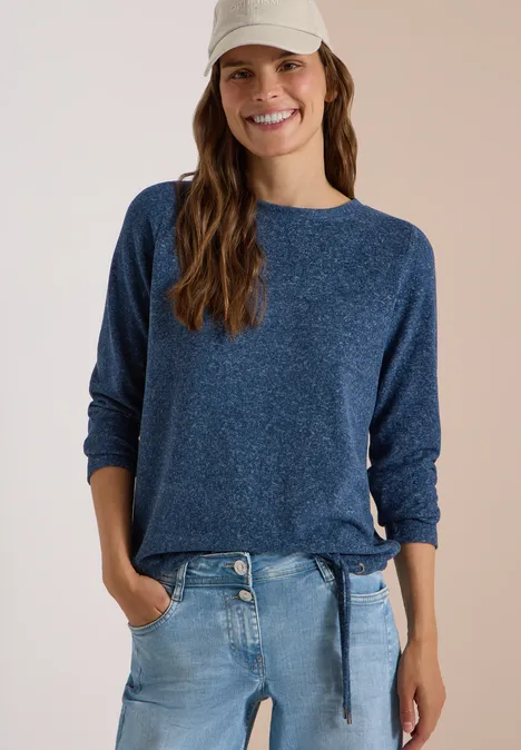 Lächelnde Frau in blauem Pullover, Jeans und Baseballcap vor hellem Hintergrund.