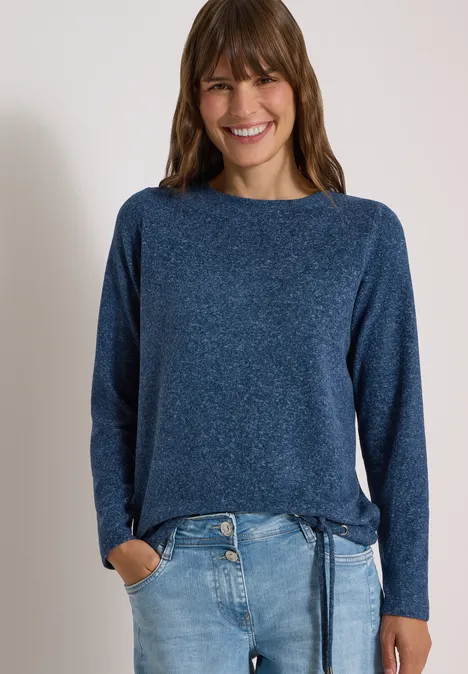 Frau mit langen braunen Haaren trägt blauen Pullover mit Bindeband und Jeans und lächelt vor hellem Hintergrund.
