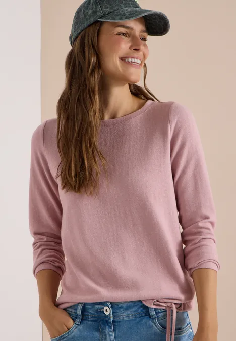 Junge Frau mit rosa Pullover und Baseball-Cap lächelt seitlich.