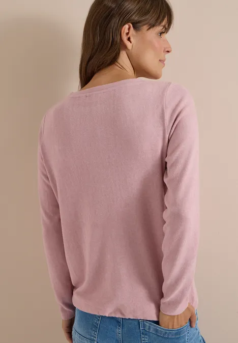 Eine Frau steht mit dem Rücken zur Kamera, trägt einen rosa Pullover und Jeans vor neutralem Hintergrund.