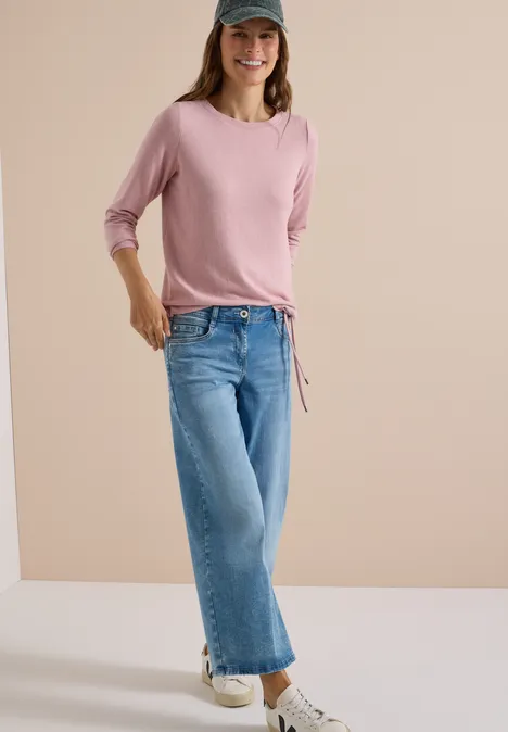 Frau in rosa Langarmshirt, Jeans, Sneakern und Baseballcap steht lächelnd mit einer Hand in der Tasche.
