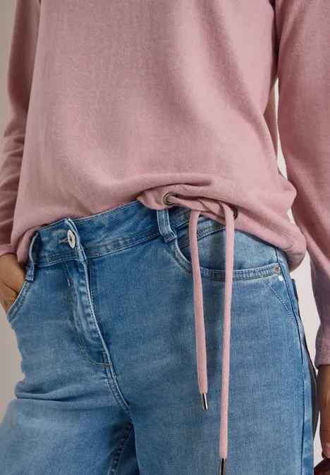 Rosafarbenes Langarmshirt mit Kordel am Saum, locker in blaue Jeans gesteckt, Hände in Taschen.