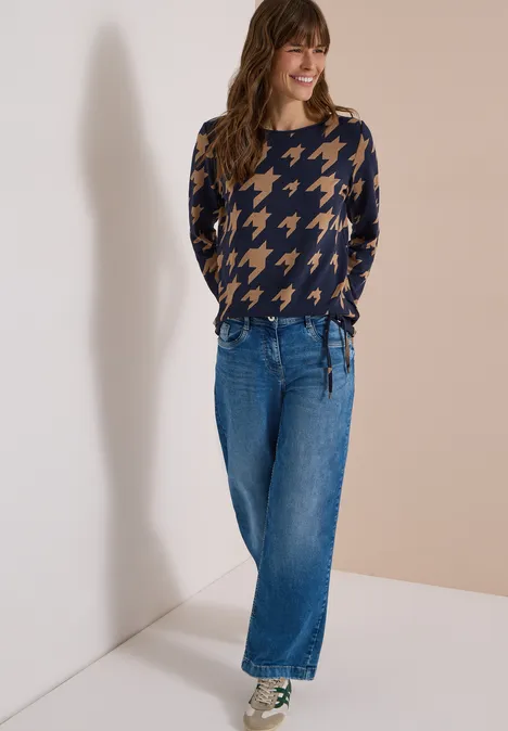 Shirt met lange mouwen, boothals en print urban dark blue Frau trägt blau-beigen Hahnentritt-Pullover und weite Jeans, steht lächelnd vor heller Wand.