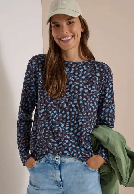 T-shirt à col bateau et motif patchwork raw chocolate brown Une femme souriante portant un haut à manches longues imprimé, un jean, une casquette et une veste verte.