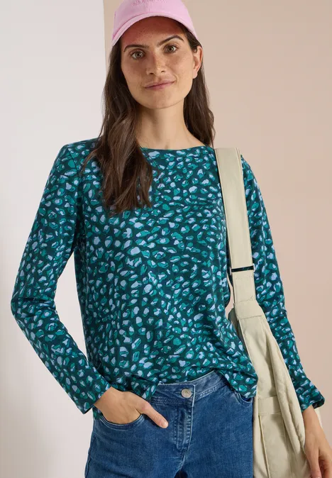 Frau in grünem gemustertem Langarmshirt und Jeans, mit Tasche über der Schulter vor hellem Hintergrund.