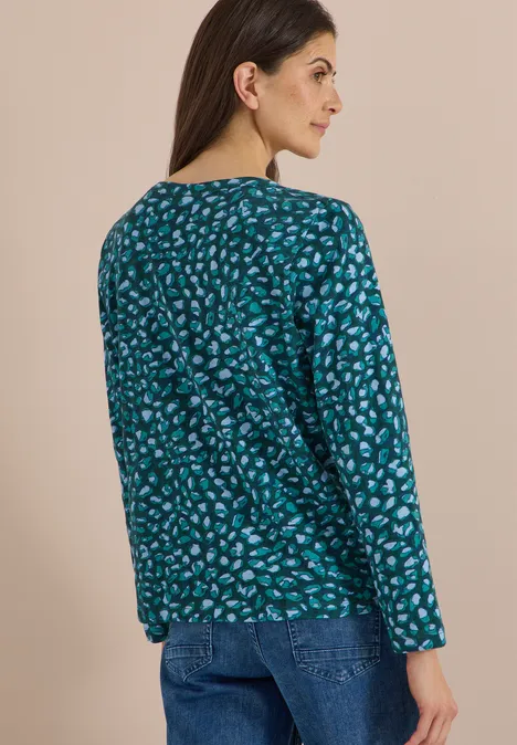 Eine Frau blickt von hinten, trägt langes braunes Haar, ein blaugrünes gemustertes Oberteil und Jeans.