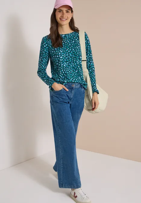 Frau in hellblauem gepunktetem Pullover, weiten Jeans, weißer Tasche und rosa Mütze lächelt vor hellem Hintergrund.