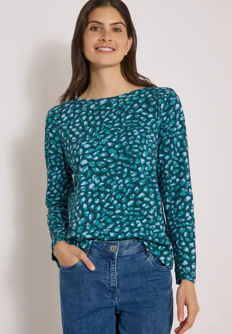 Frau in blau-grün gemustertem Langarmshirt und blauen Jeans, lächelnd, Hände in den Taschen.
