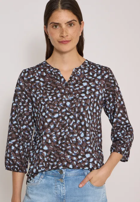 Frau in Bluse mit blauem Blumenmuster und Jeans vor hellem Hintergrund.