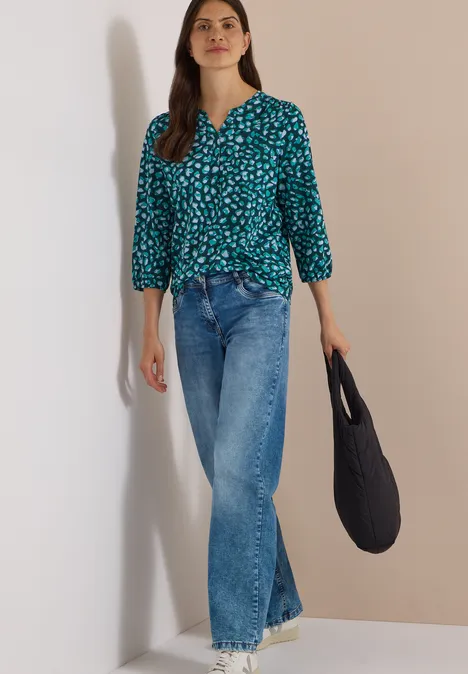 Frau in grüner Musterbluse, Jeans und schwarzer Tasche vor hellem, neutralem Hintergrund.