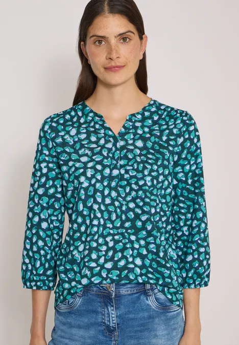 Junge Frau in blauer, gemusterter Bluse mit Dreiviertelärmeln und Jeans, Haare offen, leichter Lächeln.